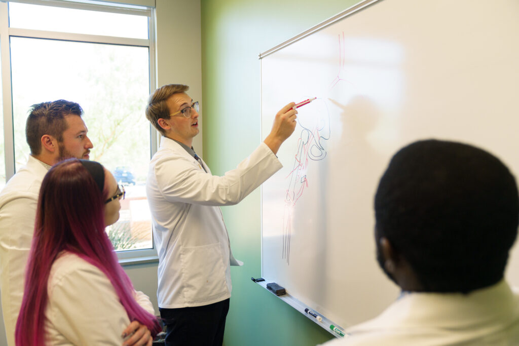 Student writing on whiteboard with classmates in white coats. 